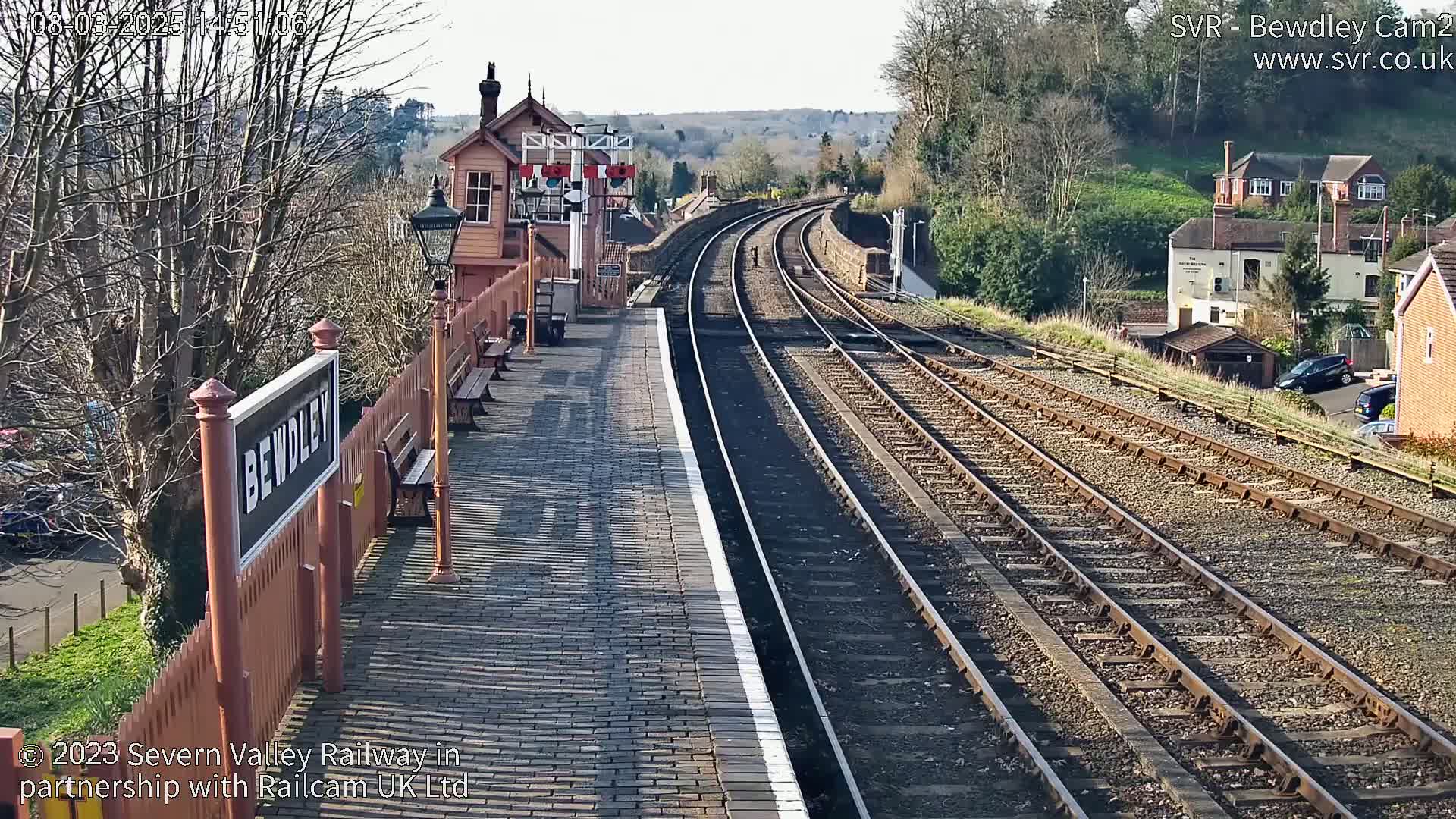A sunny view of a train station platform with multiple train tracks extending into the distance, bordered by a wooden fence and buildings.