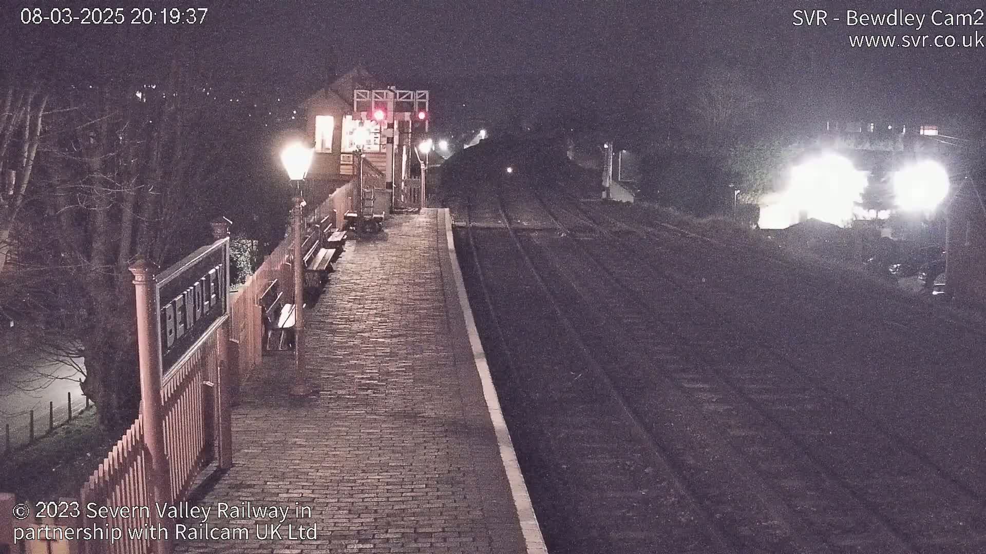 A nighttime view of a train station platform with a train approaching in the distance under dark, nighttime conditions.