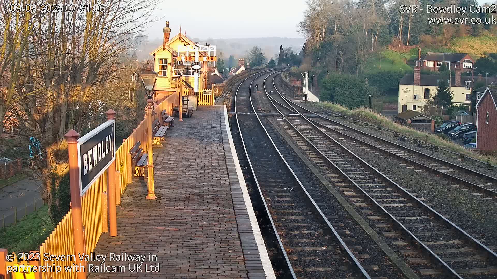 A brick train platform with a signal box and multiple train tracks stretches into the distance under a clear, bright morning sky.