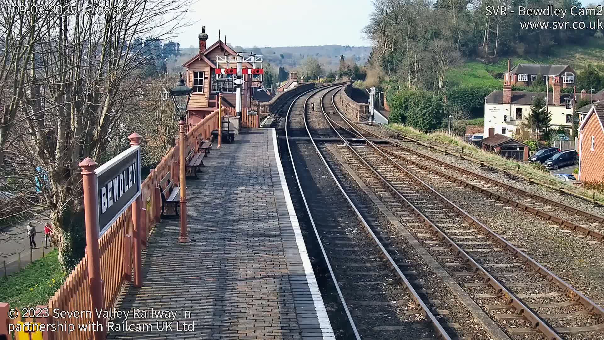 A brick railway platform with a wooden signal box and multiple train tracks extends into the distance on a sunny day.