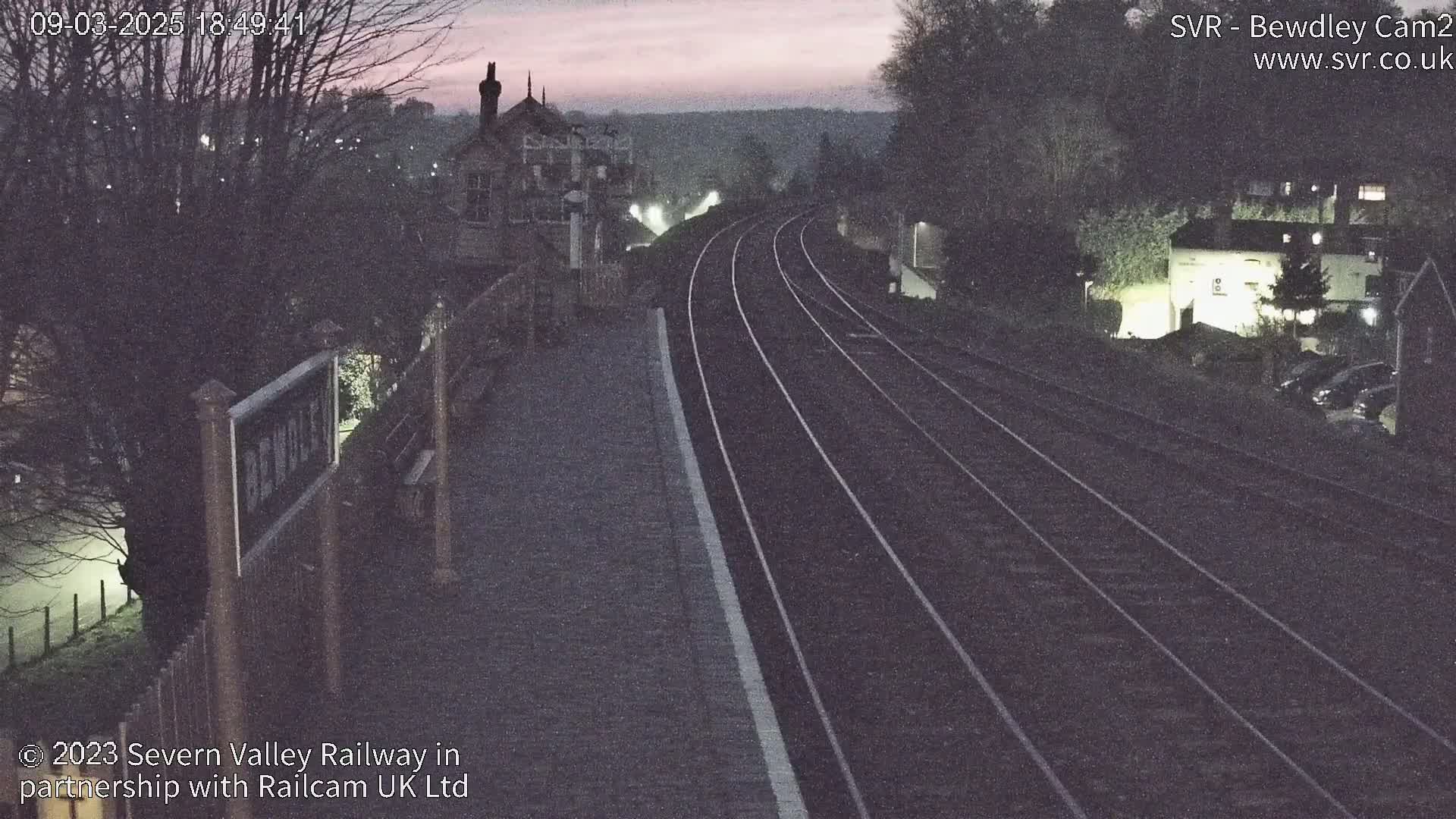 A view of a train station at twilight, showing train tracks and some buildings, under a dusky sky.