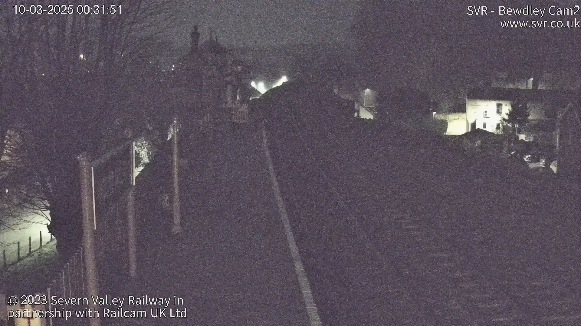 A nighttime, dimly lit view of a railway track extending into the distance, passing buildings and trees under a foggy sky.