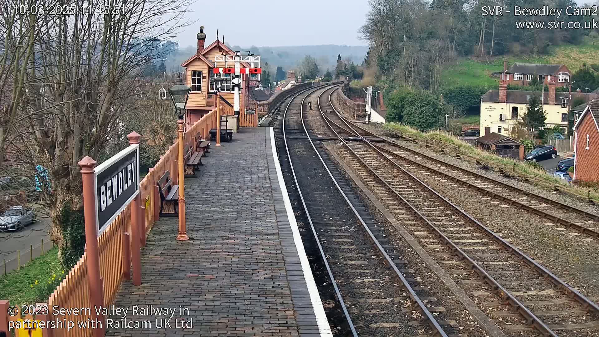 A brick train platform extends from a signal box toward curving train tracks that disappear into a hilly, residential area under an overcast sky.
