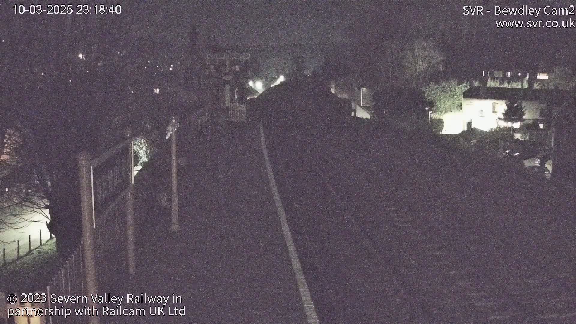 A dark, nighttime view of a railway track extending into the distance, past dimly lit buildings and trees.