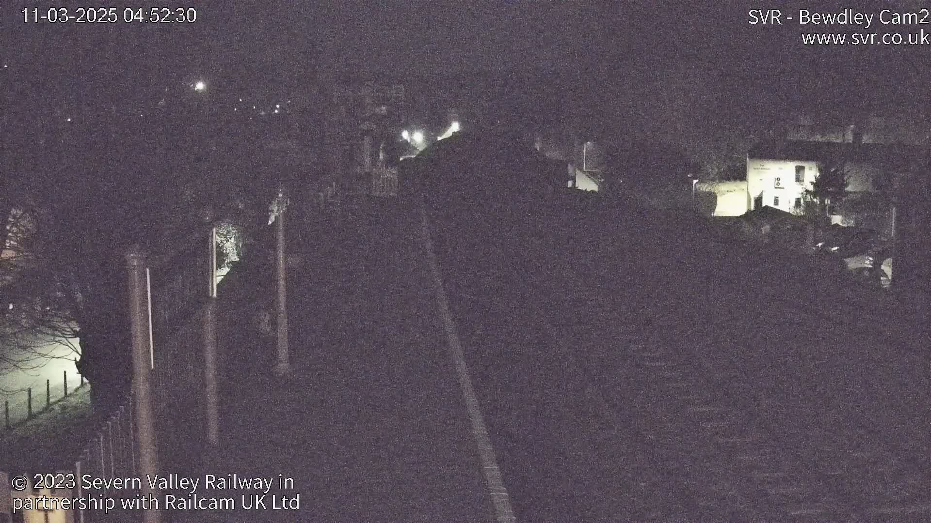 A nighttime view of a long, dark road or railway track, bordered by dark trees and buildings, under a clear, dark sky.
