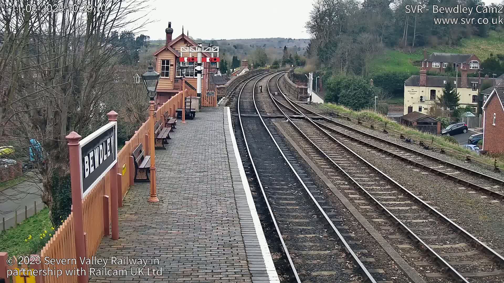 A railway station platform with brick paving, a signal box, and multiple train tracks extending into the distance under an overcast sky.