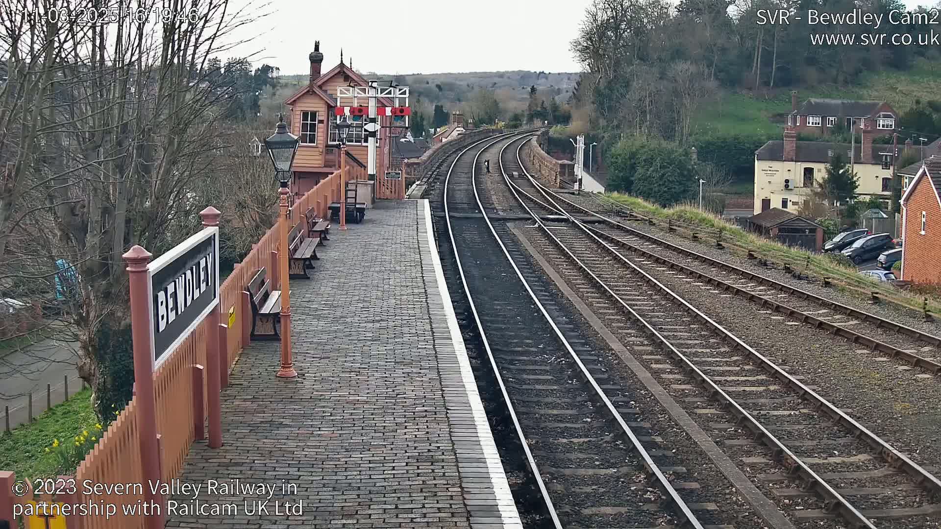 A brick railway platform with a station building and multiple train tracks stretches into the distance on an overcast day.