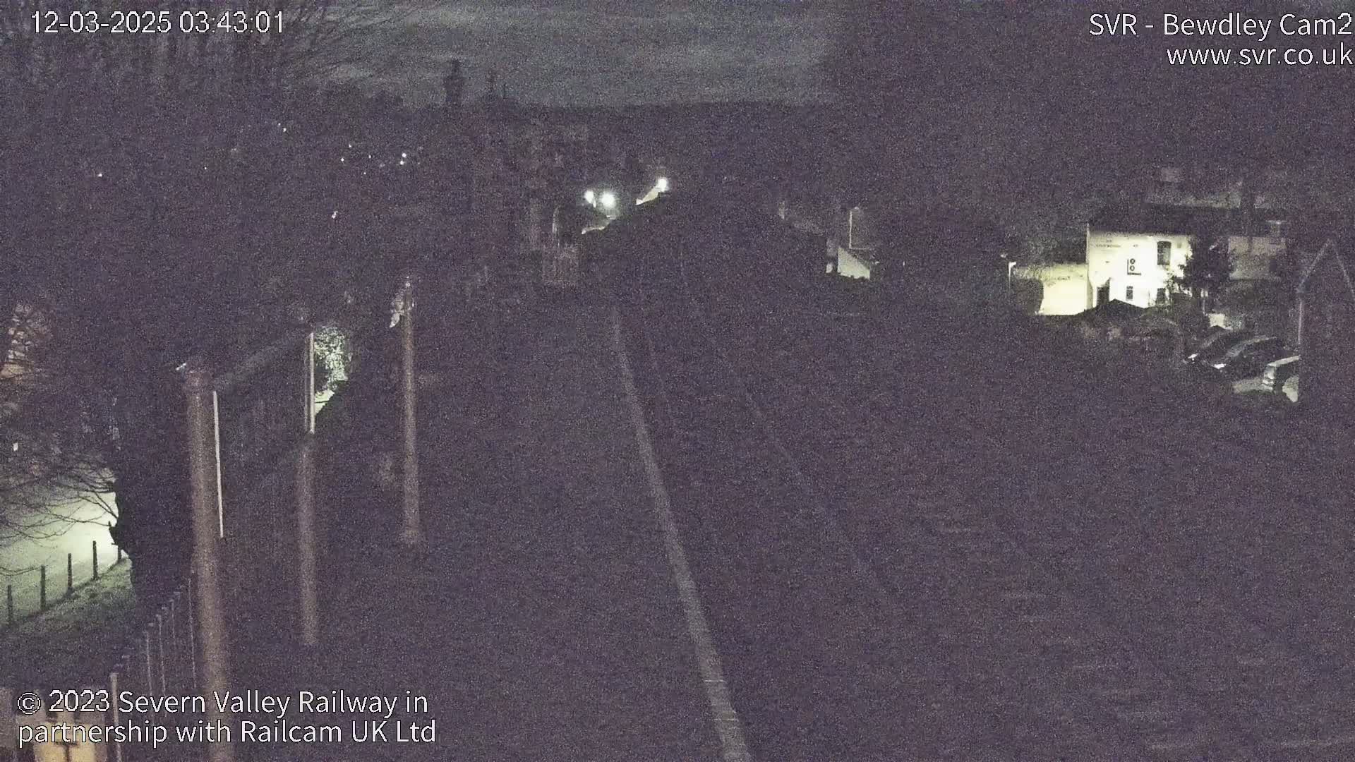 A dark, nighttime view of a railway track, bordered by trees and buildings with dim lighting.