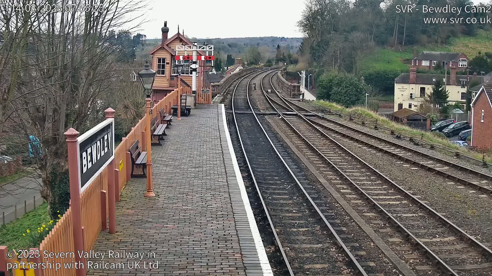 A train station platform with brick paving and wooden benches overlooks curving railway tracks on an overcast day.
