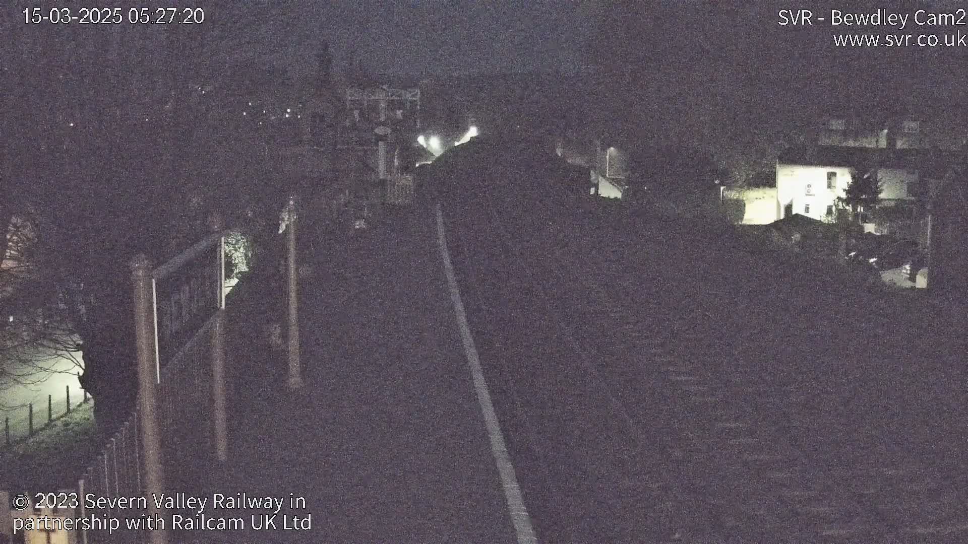 A nighttime view of a railway track stretching into the distance, with dimly lit buildings and trees visible in the background under dark, possibly foggy conditions.