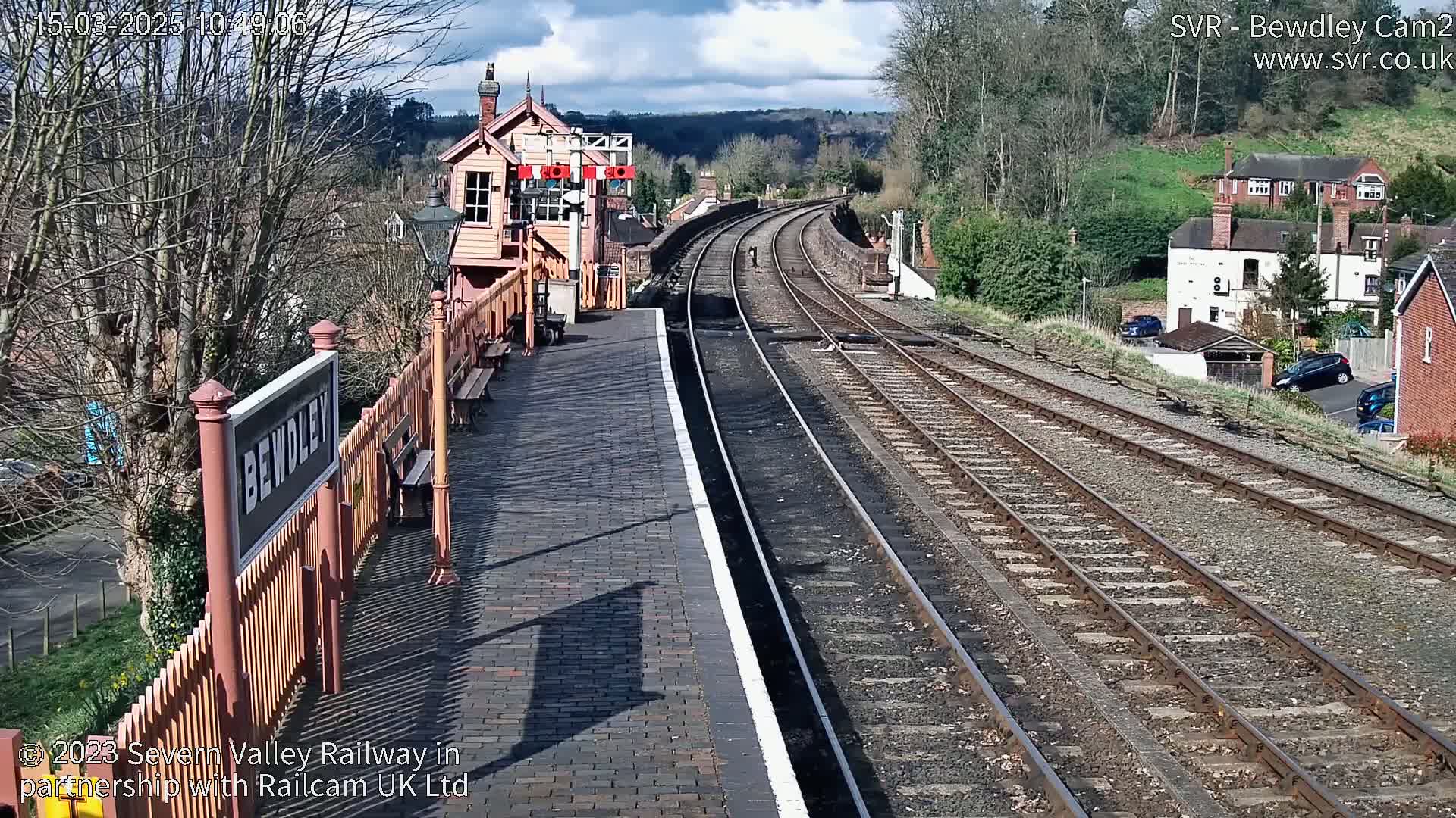A sunny view of a railway station platform with curving train tracks extending into the distance, beyond which are houses and hills.