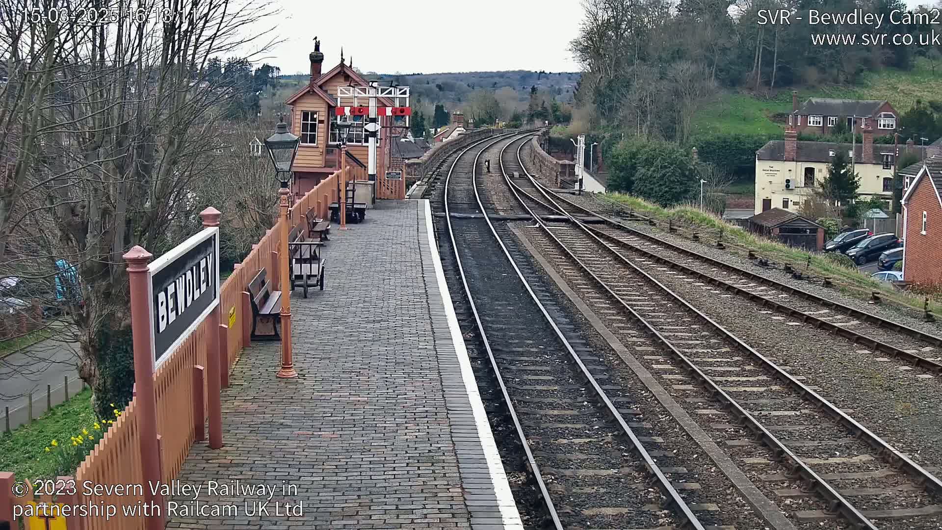 A train station platform with a brick pavement and multiple train tracks stretches into the distance under an overcast sky.