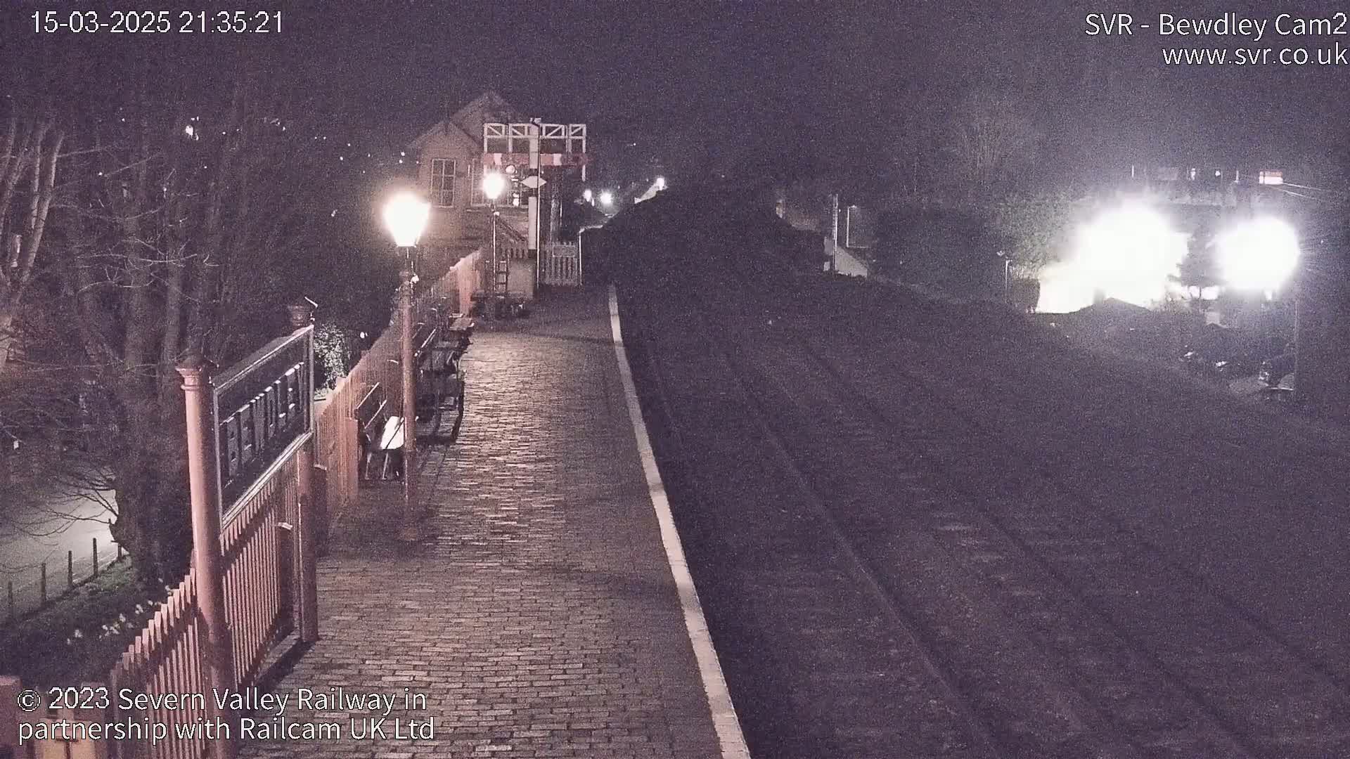 A nighttime view of a train platform with a brick pathway, a gate, and a train approaching in the distance under dark skies.