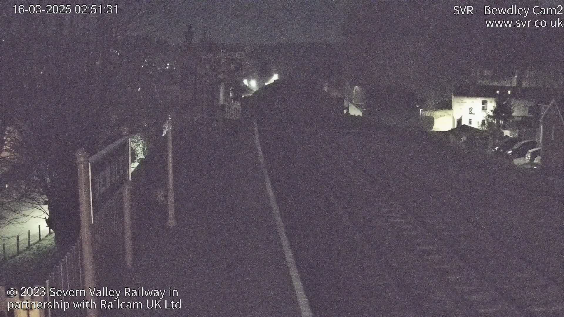 A nighttime view of a dimly lit road or railway track, flanked by dark buildings and trees under a dark sky.