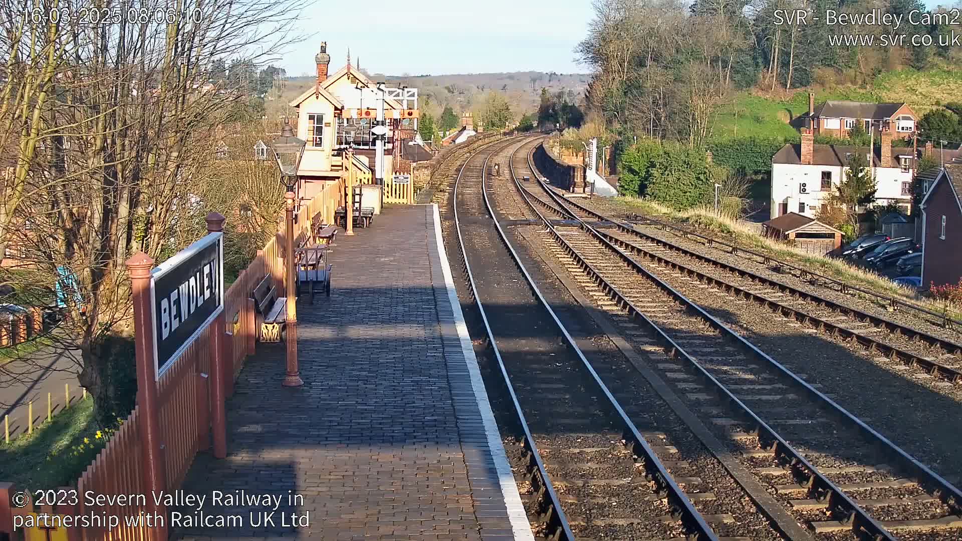 A sunny view of a train station platform with tracks extending into the distance, surrounded by buildings and trees.