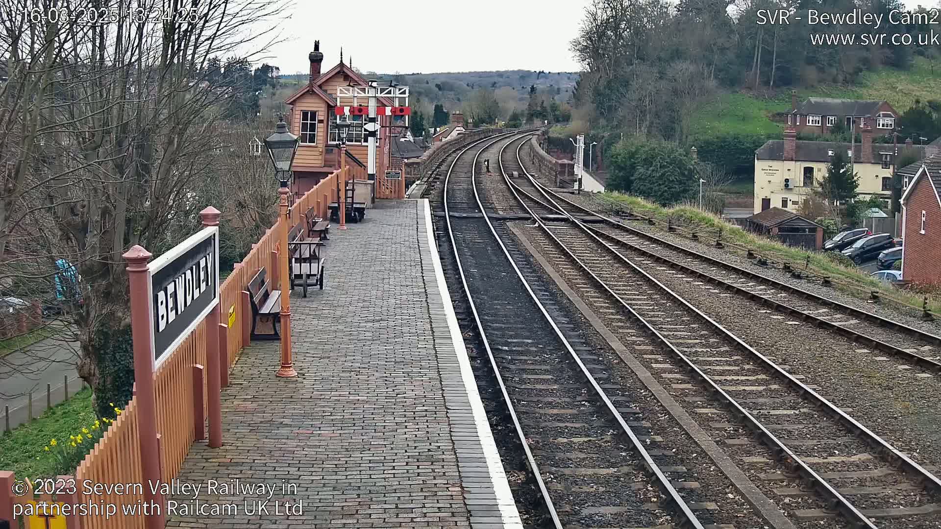 A brick railway platform with a signal box and multiple train tracks extends into the distance under an overcast sky.