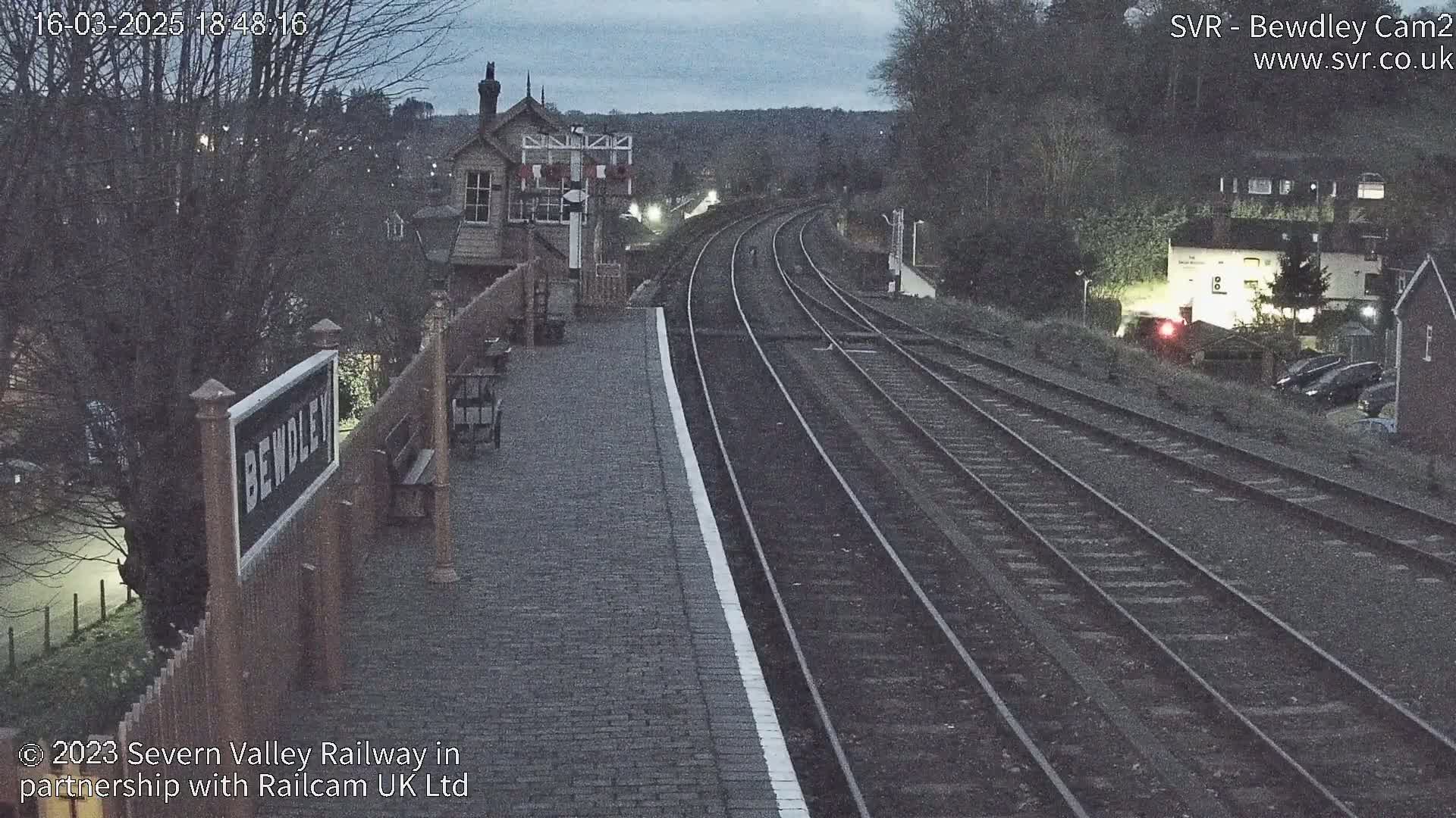 A train station platform at dusk shows multiple train tracks extending into the distance under a dark, overcast sky.