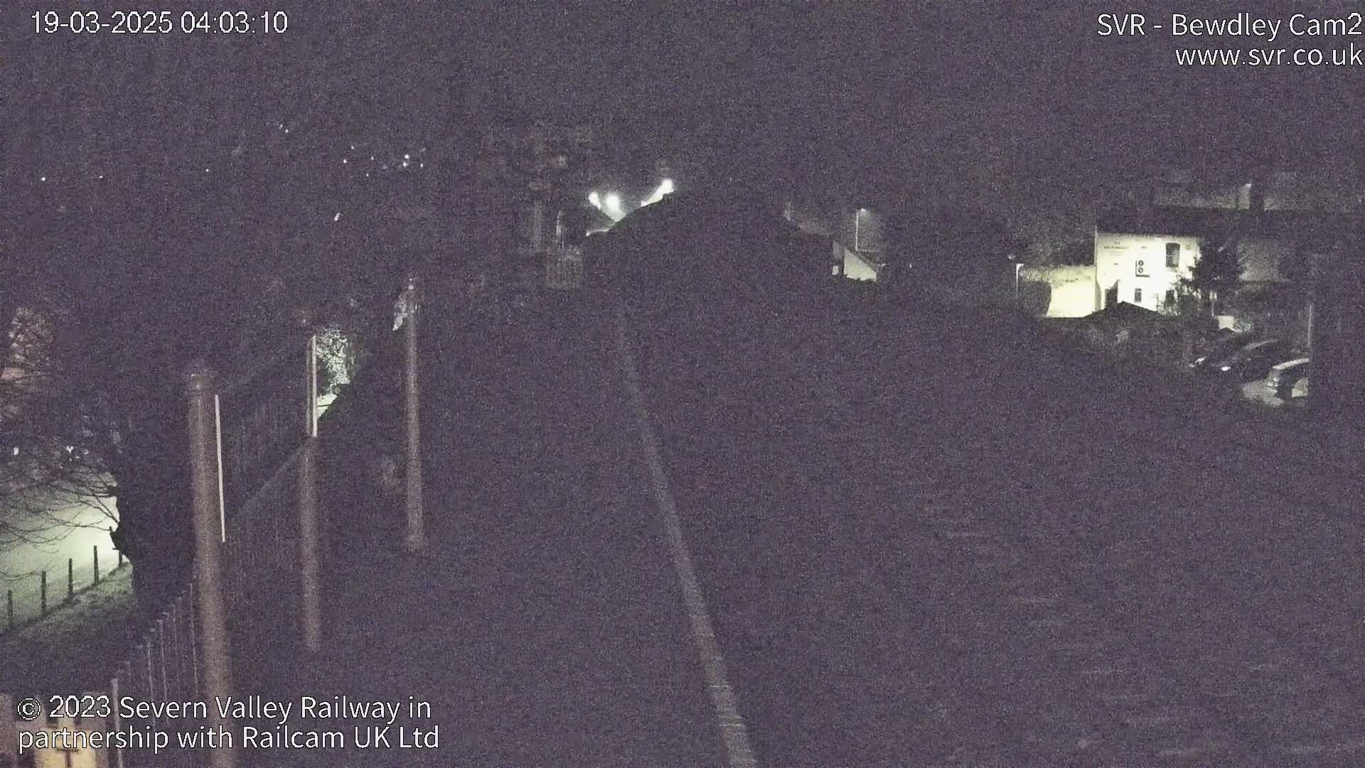 A nighttime, low-light view shows a railway track leading toward a dimly lit building and trees.