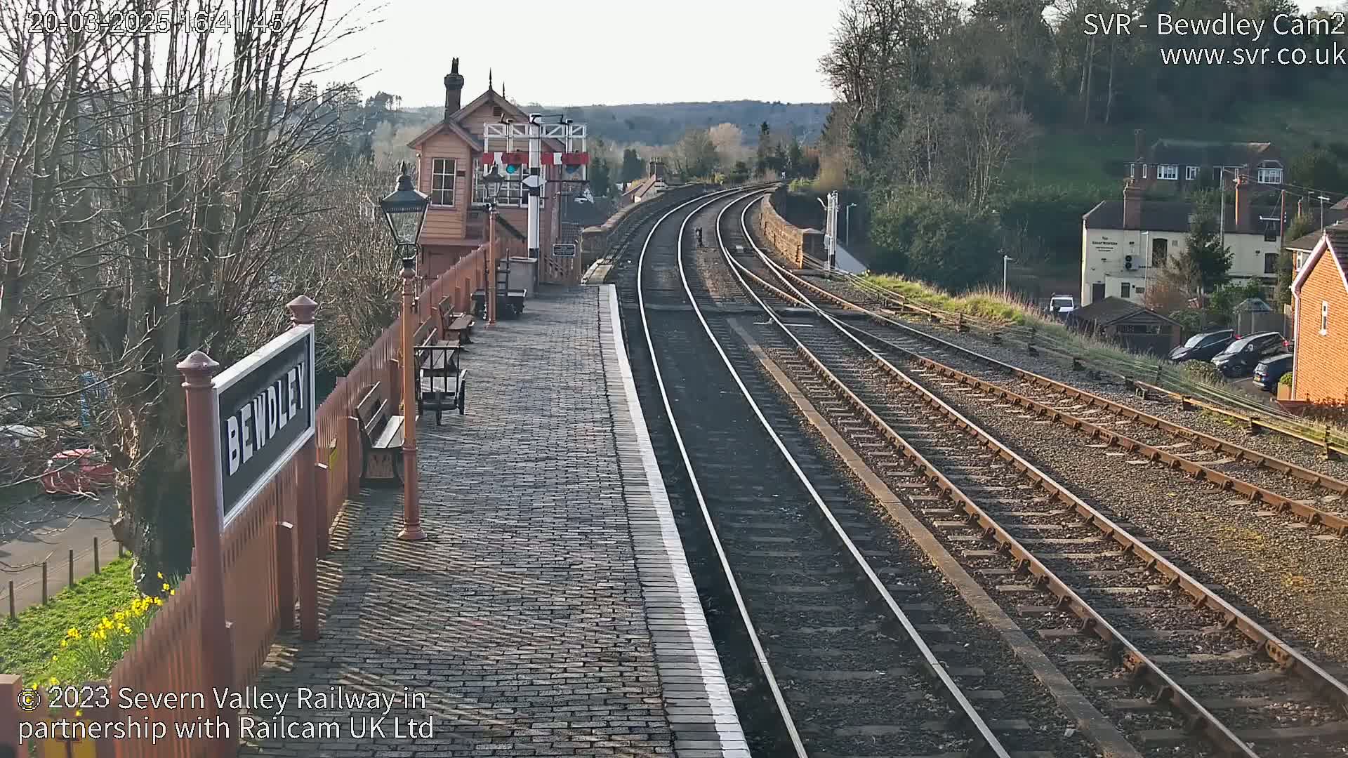 A train station platform with a brick paved surface, multiple train tracks extending into the distance, a signal box, and some parked cars visible beyond the tracks on a sunny day.