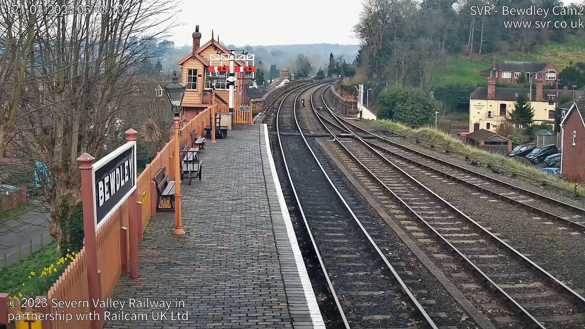 A train station platform with a brick surface and multiple train tracks extends into the distance under an overcast sky, bordered by buildings and trees.