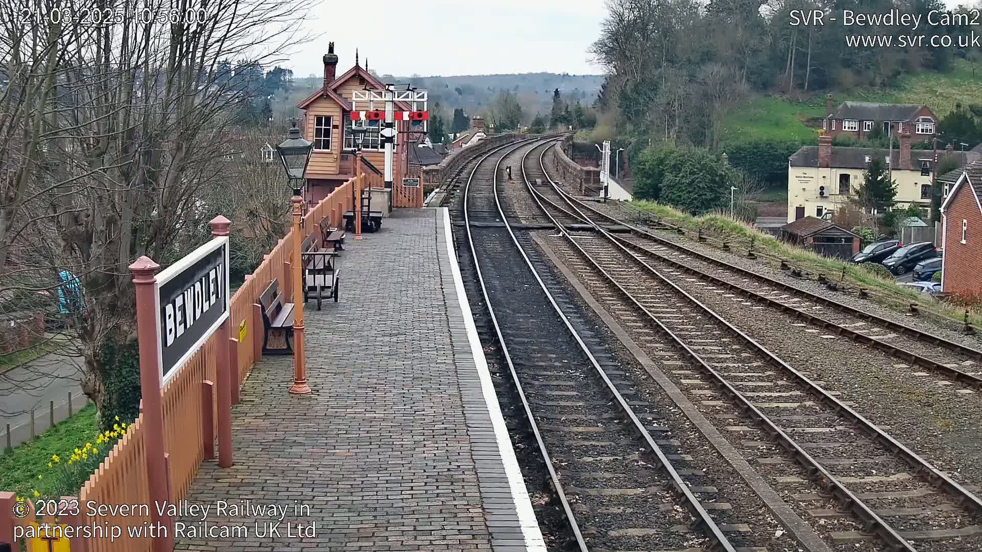 A train station platform with a brick surface and wooden benches, overlooking several train tracks that curve into the distance under a cloudy sky.