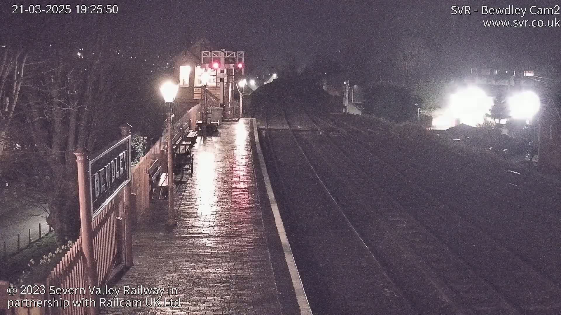 A wet, empty train platform at night is illuminated by streetlights, with train tracks extending into the darkness.