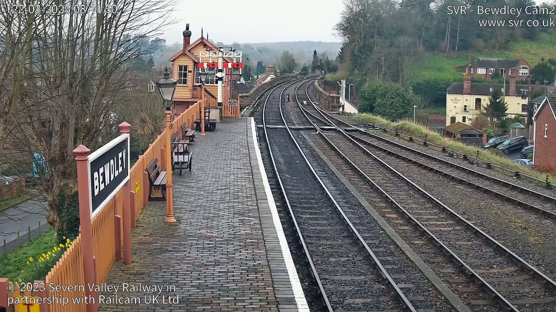 A train station platform with multiple train tracks stretches into the distance under an overcast sky, with a small building and houses visible in the background.