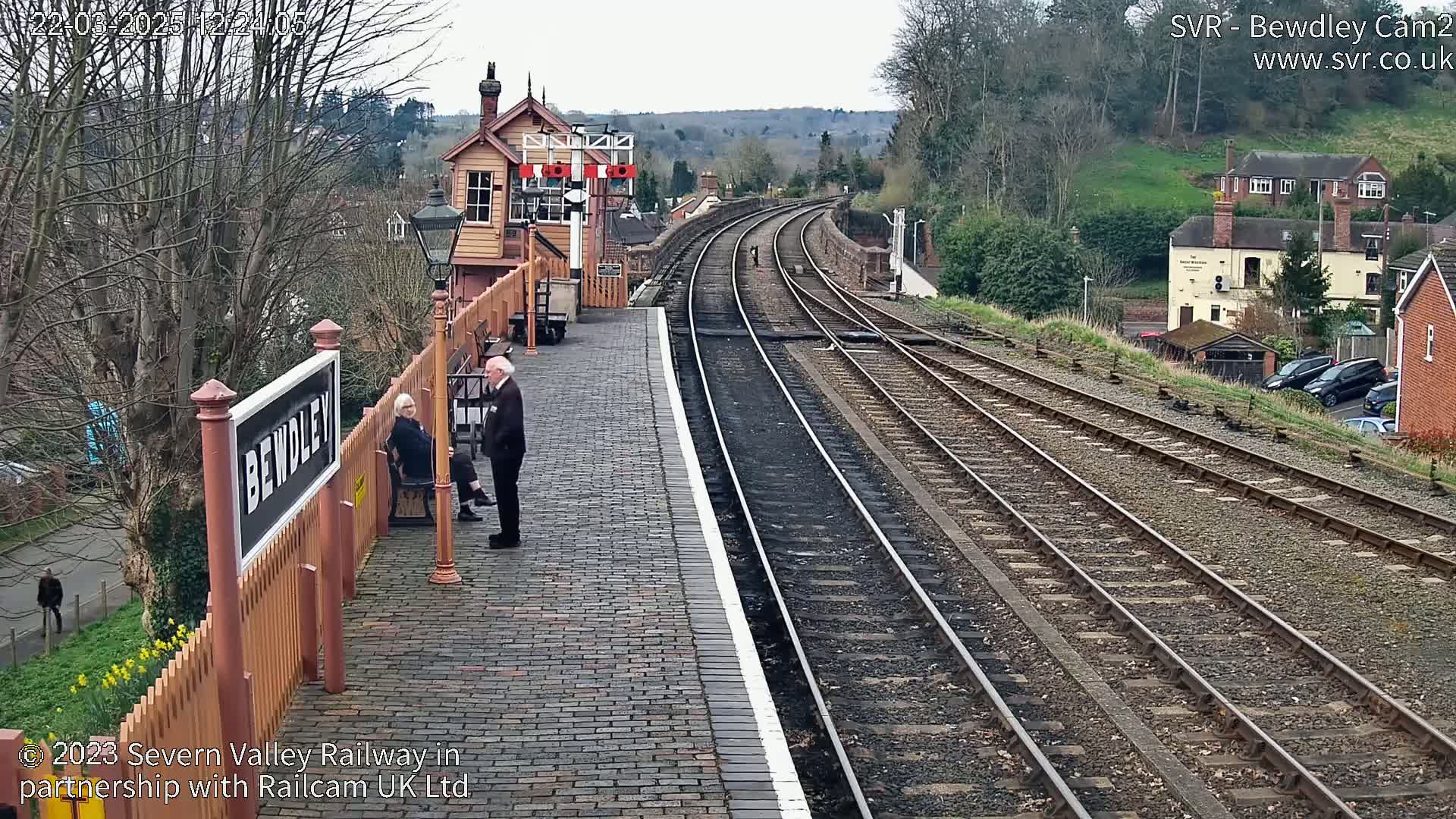 A brick train platform with two people standing near a sign, overlooking several train tracks that curve into the distance under a partly cloudy sky.