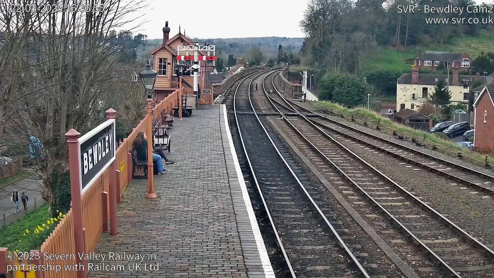 A train station platform with two people sitting on a bench, overlooking a railway track that curves into the distance under an overcast sky.