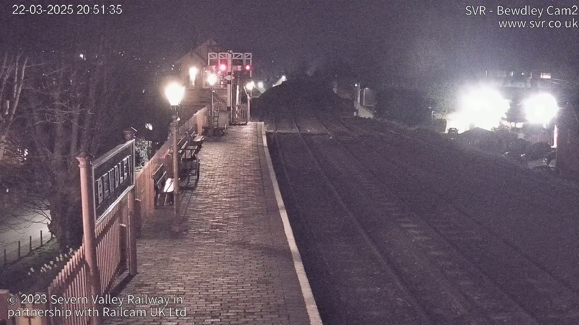 A nighttime view of a brick railway platform, illuminated by lampposts, with train tracks extending into the darkness.
