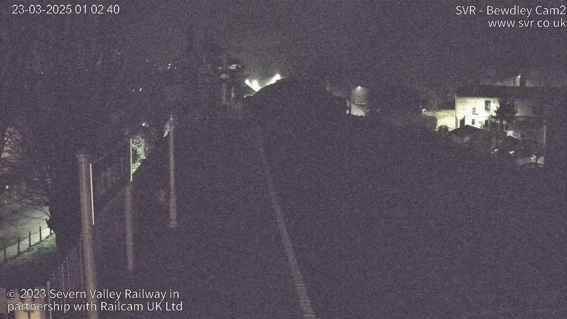 A nighttime, low-light view of a dimly lit area, possibly a railway yard, with some buildings visible in the distance under a dark sky.