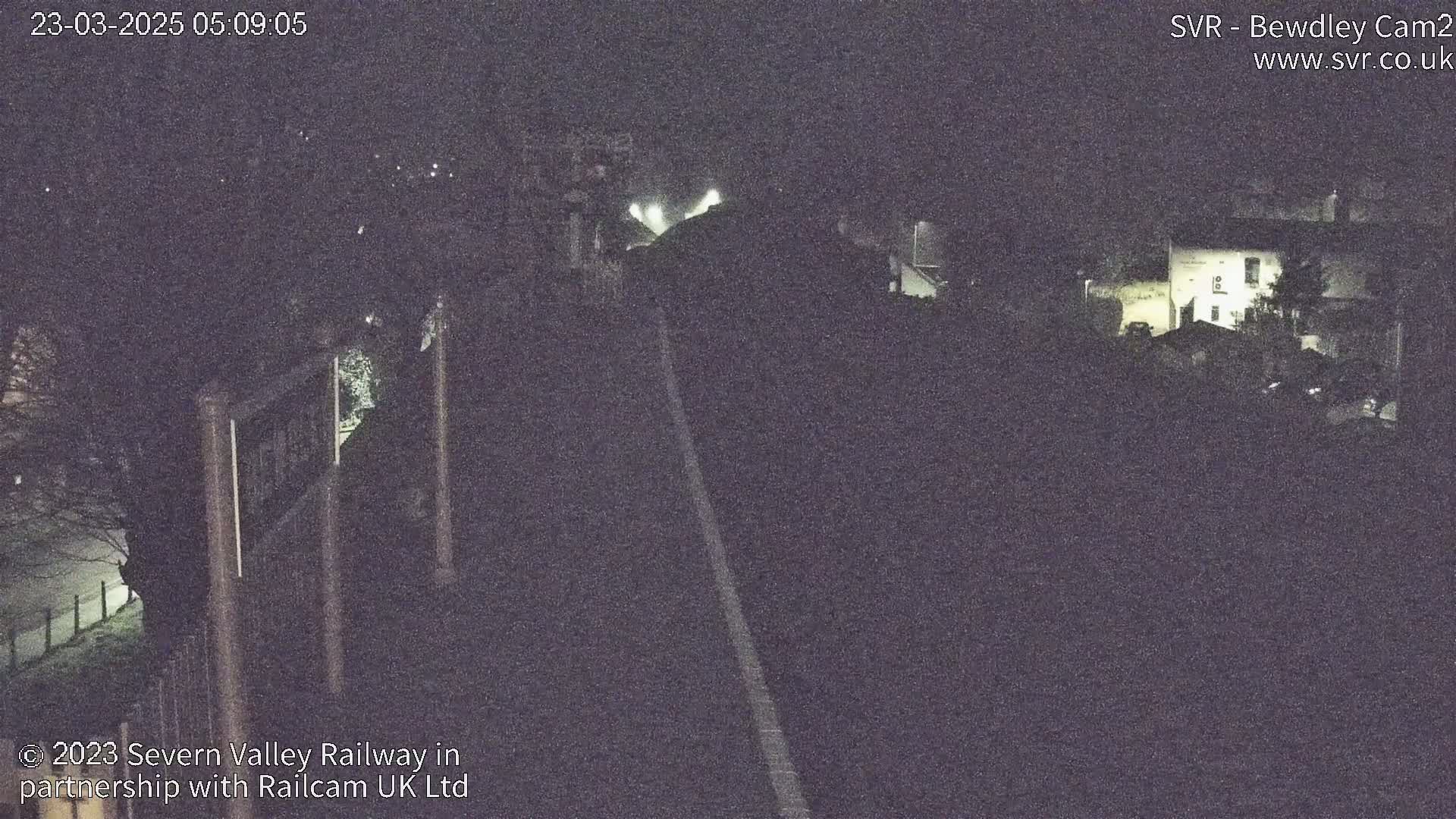 A nighttime, low-light view of a railway track extending toward a dimly lit building and trees.