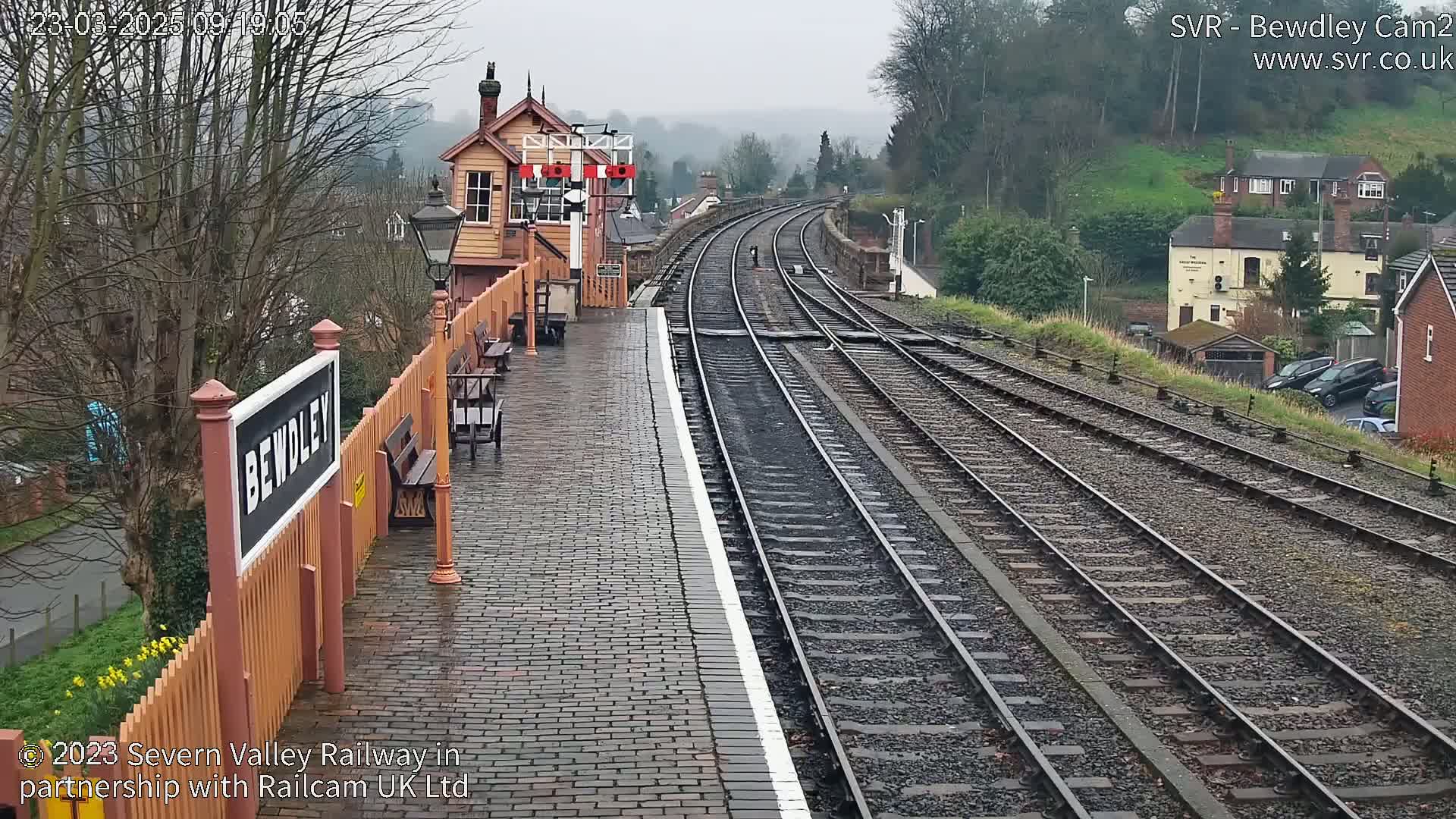 A wet, overcast day at a train station shows a brick platform next to several train tracks curving into the distance, with a signal box and buildings visible in the background.