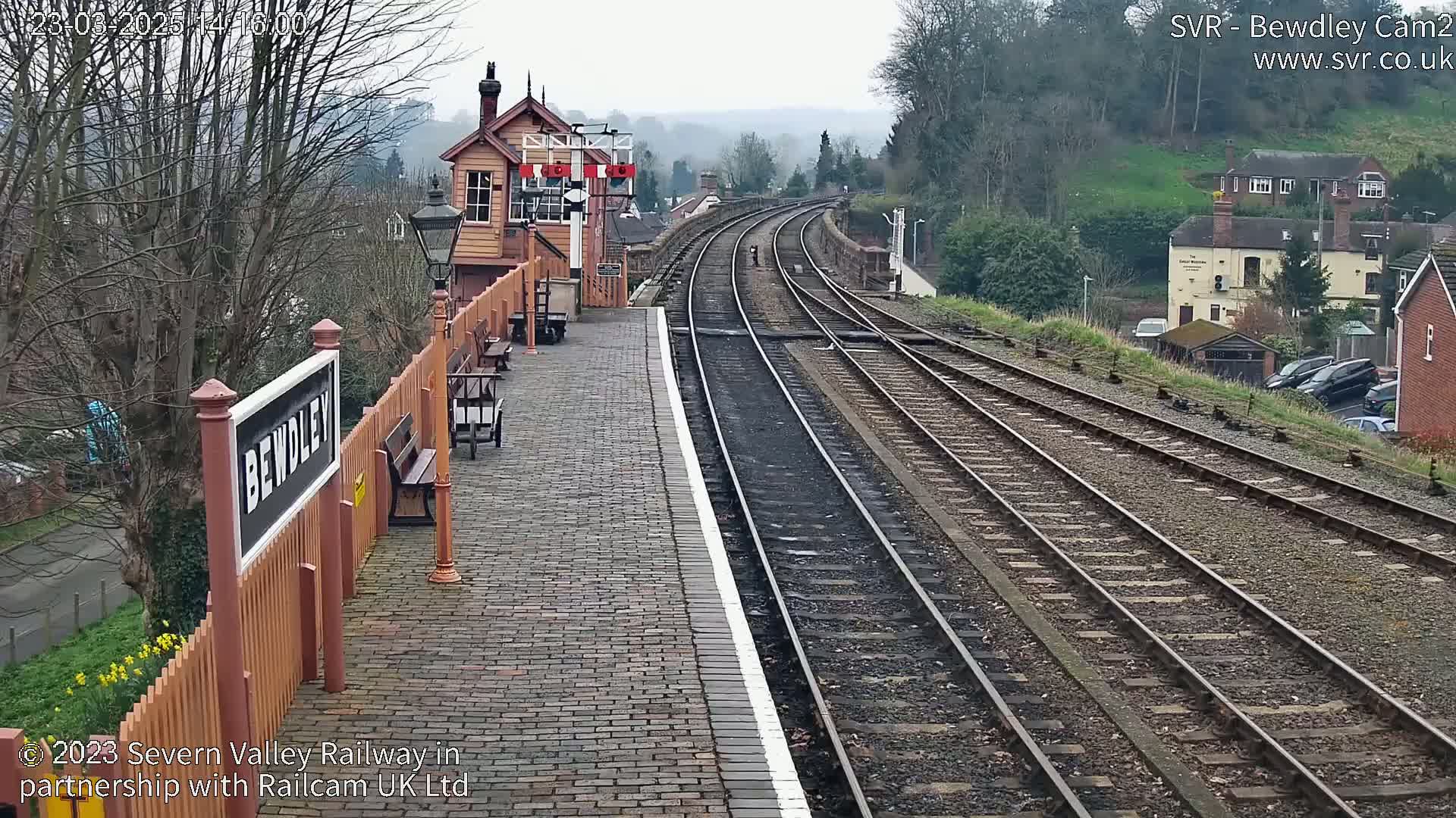 A train station platform with multiple train tracks extending into the distance on an overcast day, featuring a signal box and buildings in the background.