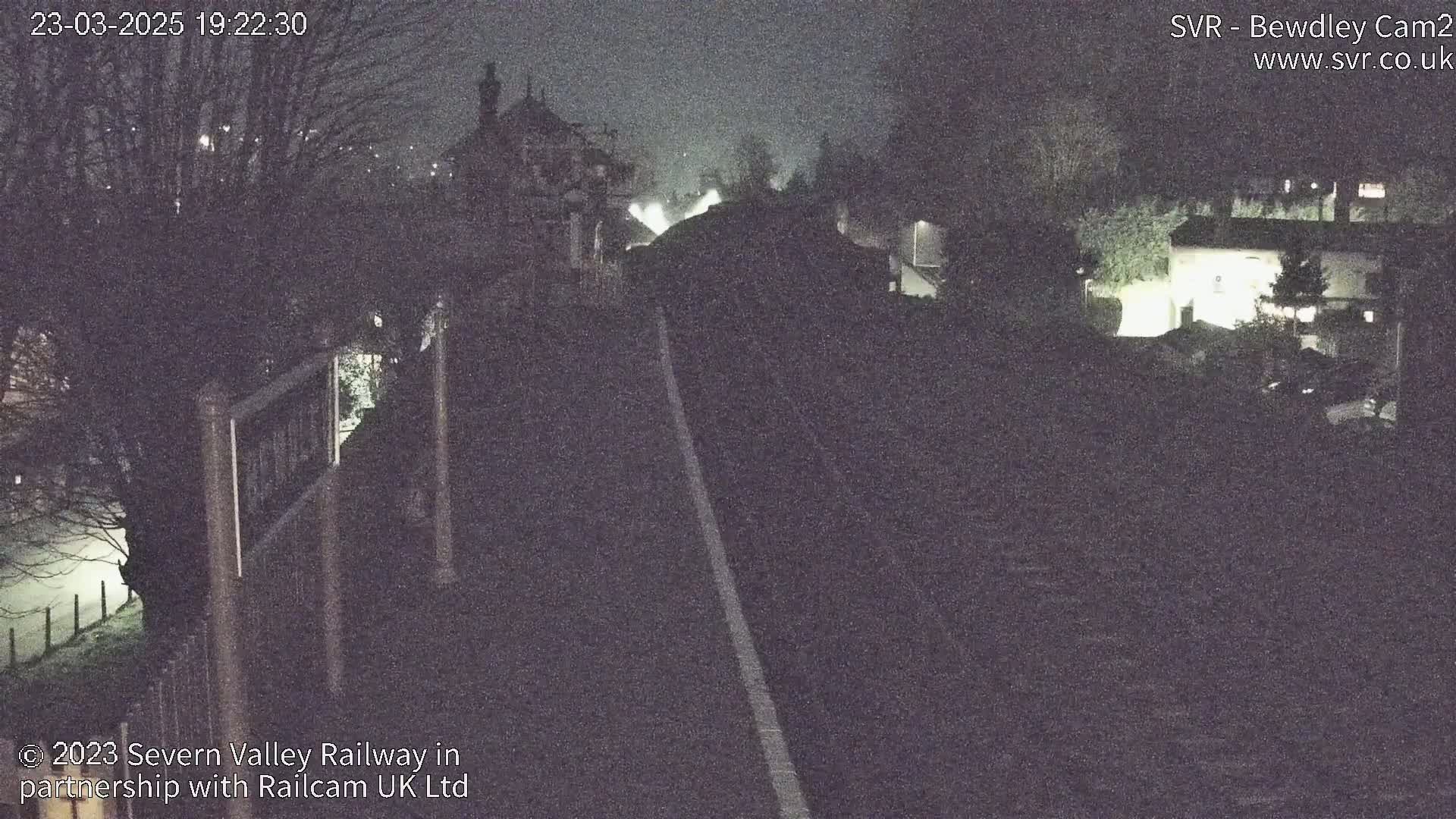 A nighttime view of a train track, bordered by trees and buildings, under foggy or misty conditions.