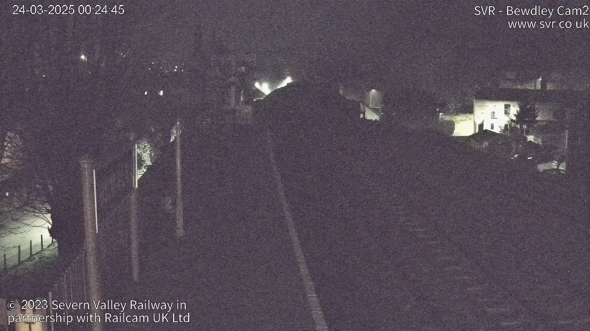 A nighttime, low-light view of a railway track leading toward some buildings and trees, with a foggy or misty atmosphere present.