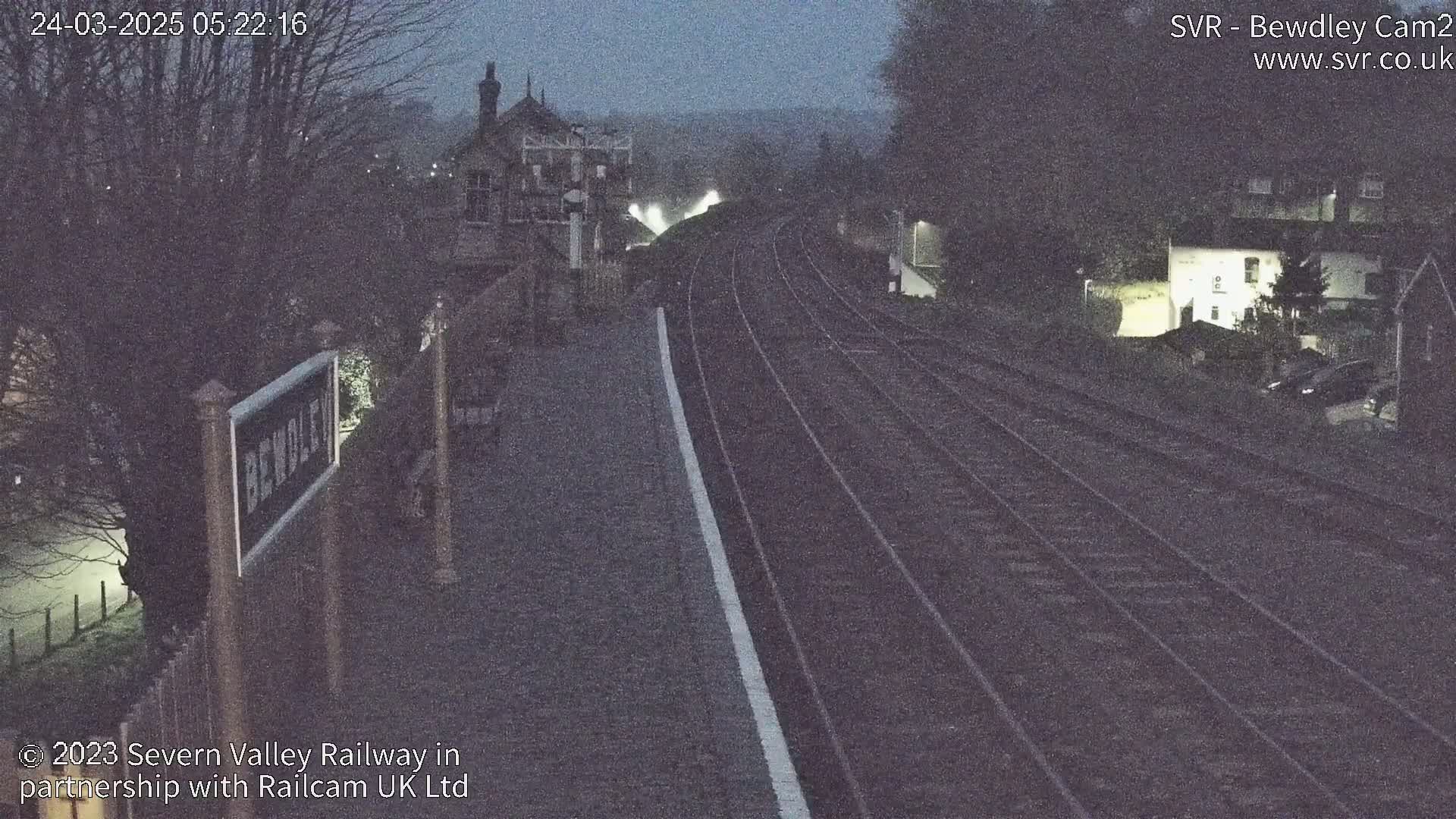 A dark, nighttime view of train tracks extending into the distance, with a station building and surrounding structures visible under a dim sky.