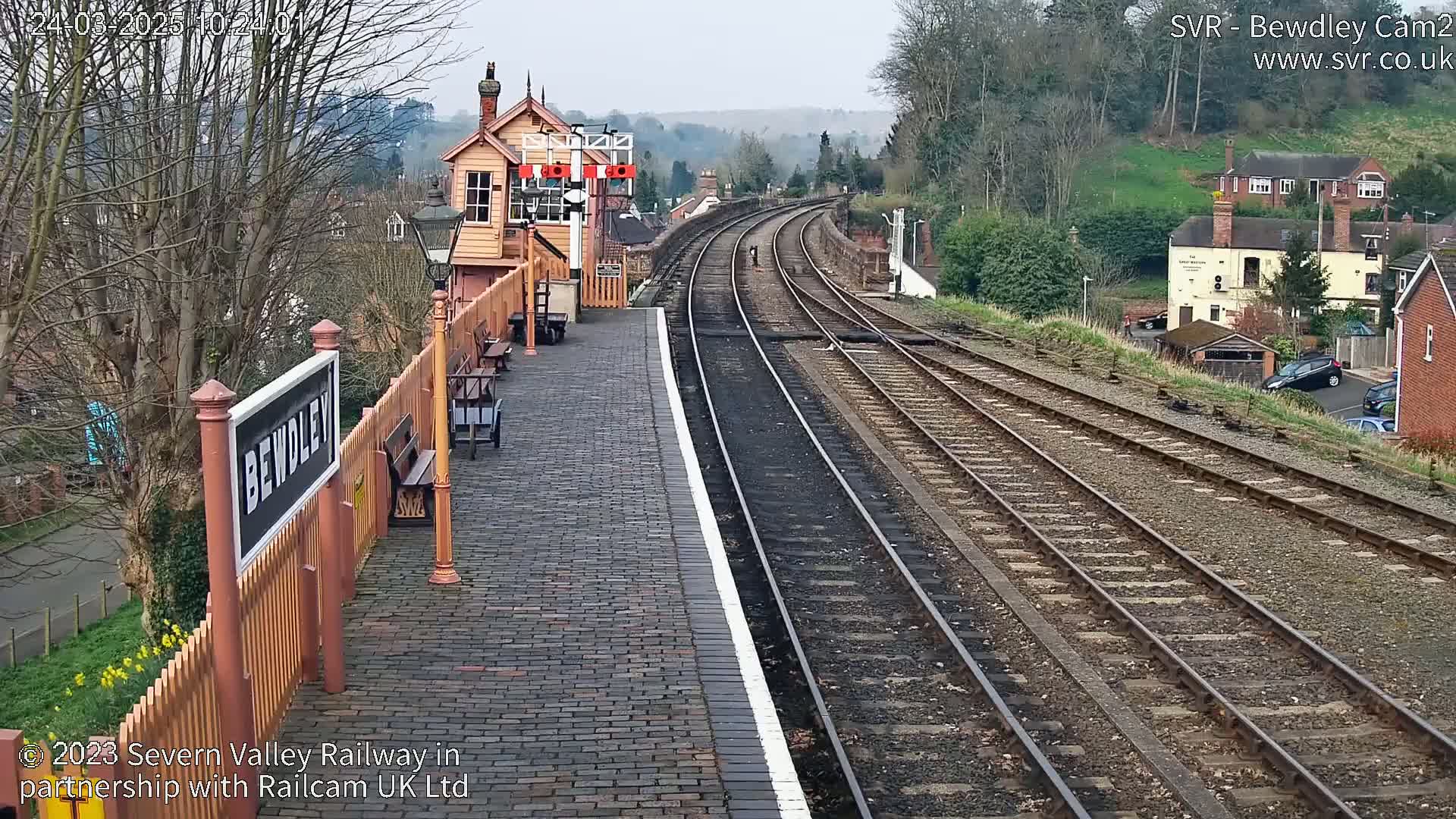A brick railway platform with a signal box and curving tracks extends into a tree-lined valley under a cloudy sky.