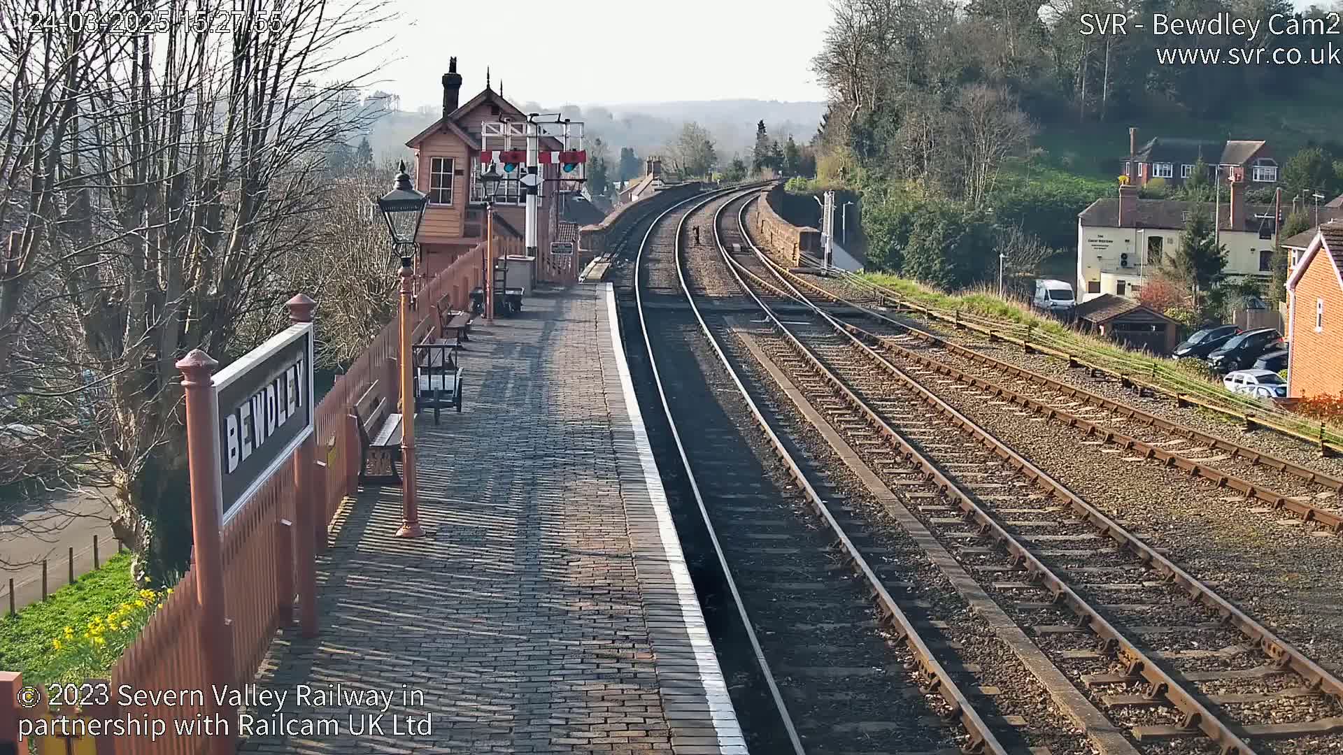 A sunny view of a train station platform with multiple train tracks extending into the distance, beside a residential area.