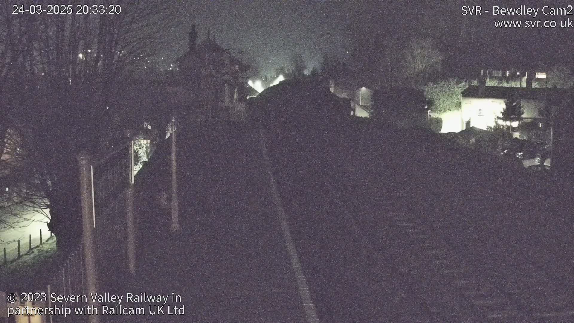 A nighttime, low-visibility view of a railway track passing through a dimly lit area with buildings and trees on either side.