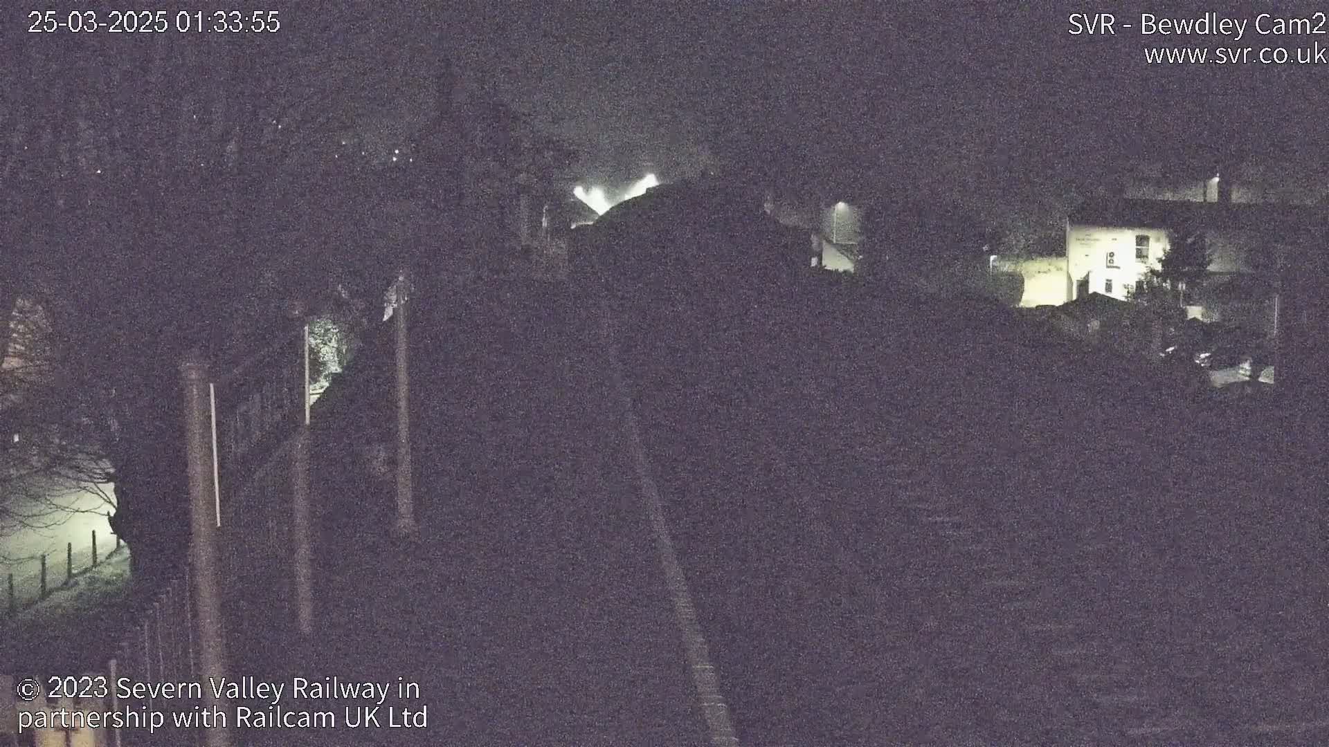 A nighttime, low-light view of a railway line and buildings, possibly a station, under overcast conditions.