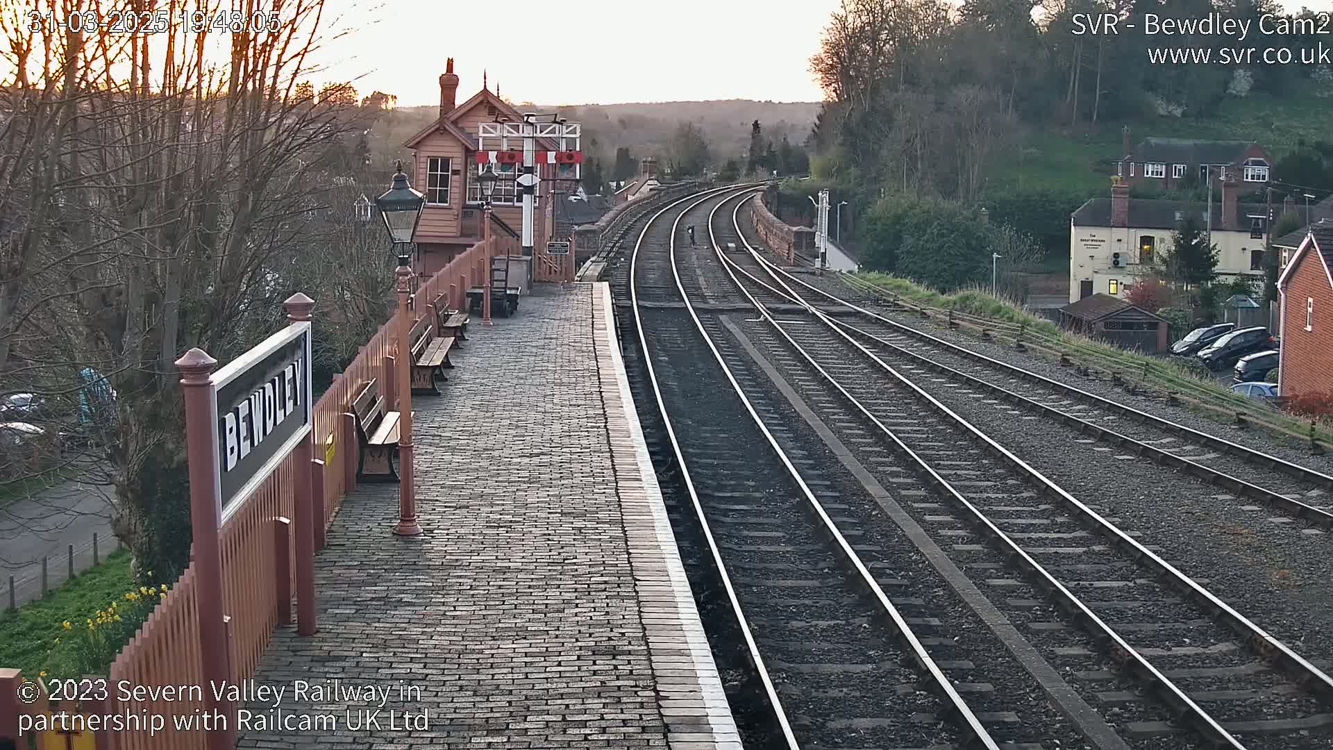 A train station platform with several train tracks extends into the distance under a partly cloudy evening sky.