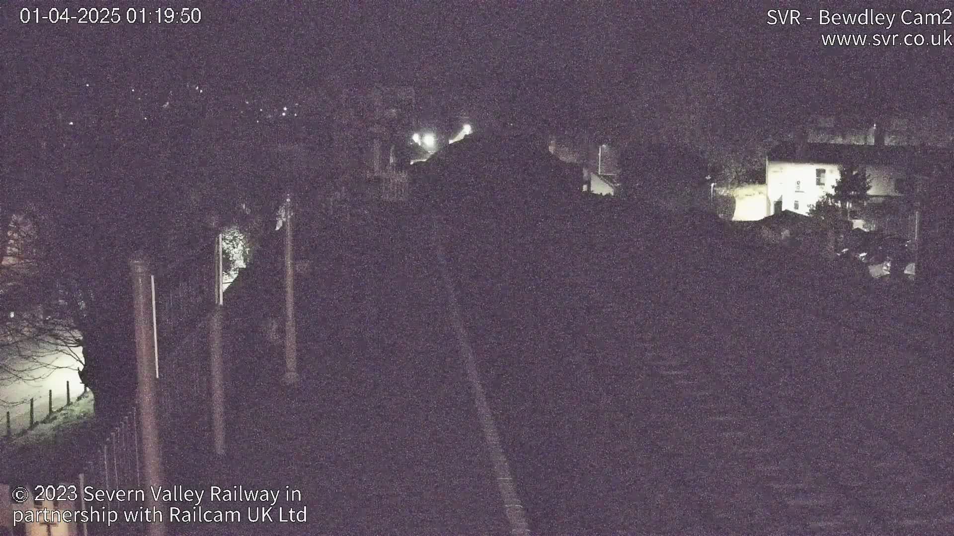 A nighttime, low-light view shows a railway track leading towards a building and trees under a dark sky.