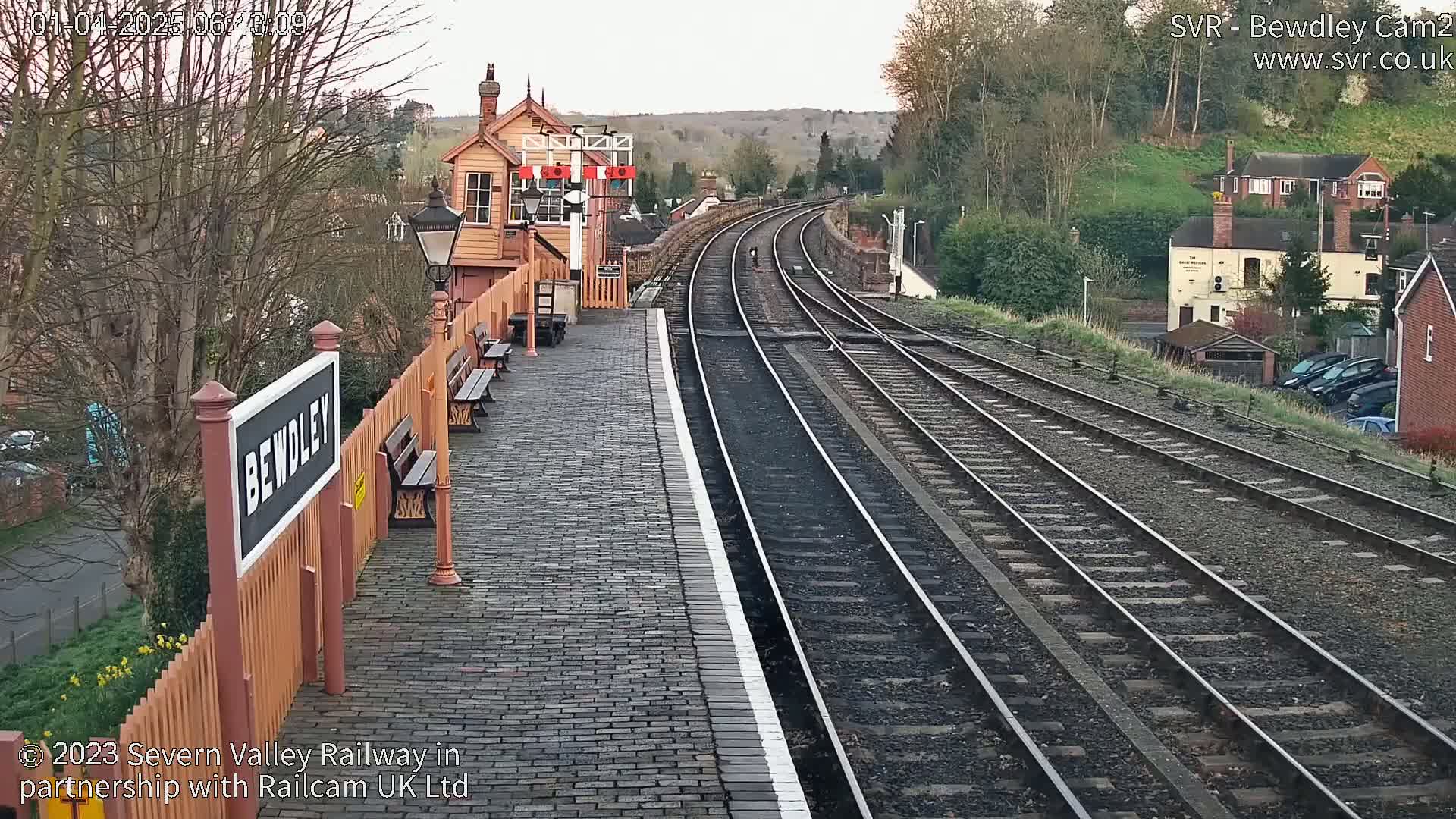 A railway station platform with a brick surface and wooden benches, overlooking several train tracks that curve into the distance under a partly cloudy sky.