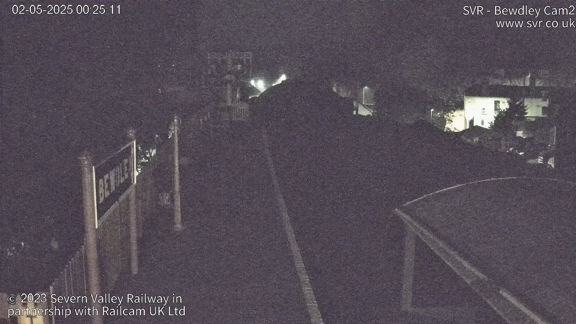 A nighttime, low-light view of a train platform with a partially visible station sign and buildings in the background.