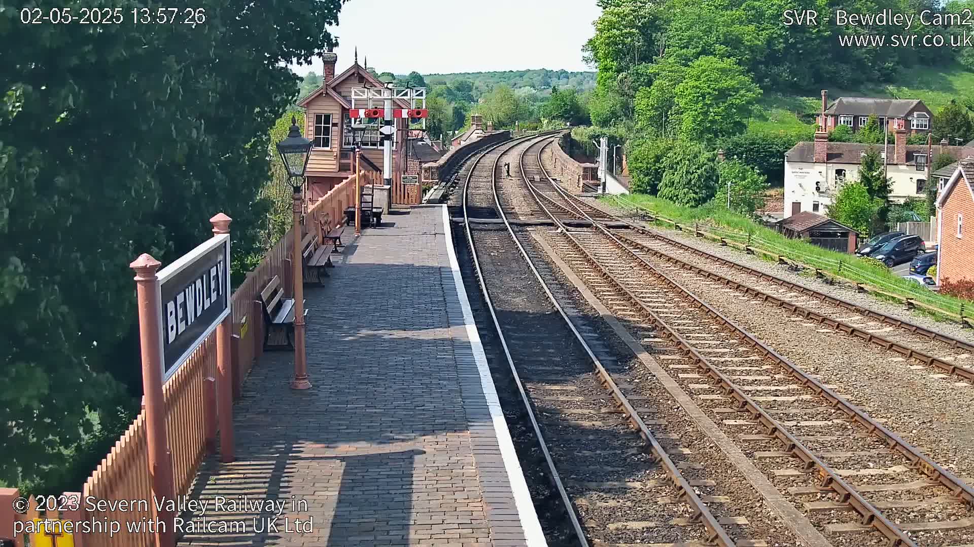 A sunny view of a railway station platform and tracks curving into the distance, with a signal box and houses visible beyond the tracks.