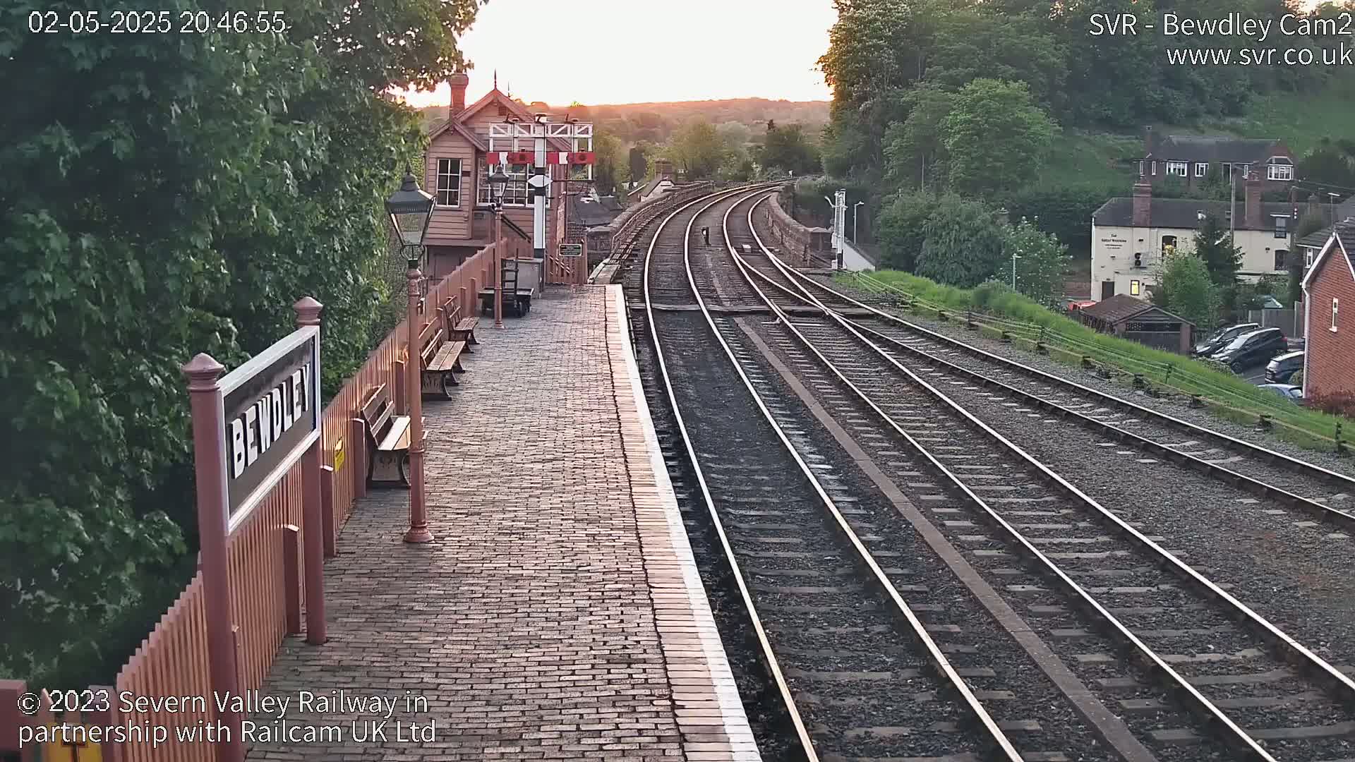 A brick railway platform at sunset, with multiple train tracks extending into the distance, flanked by lush greenery and buildings.