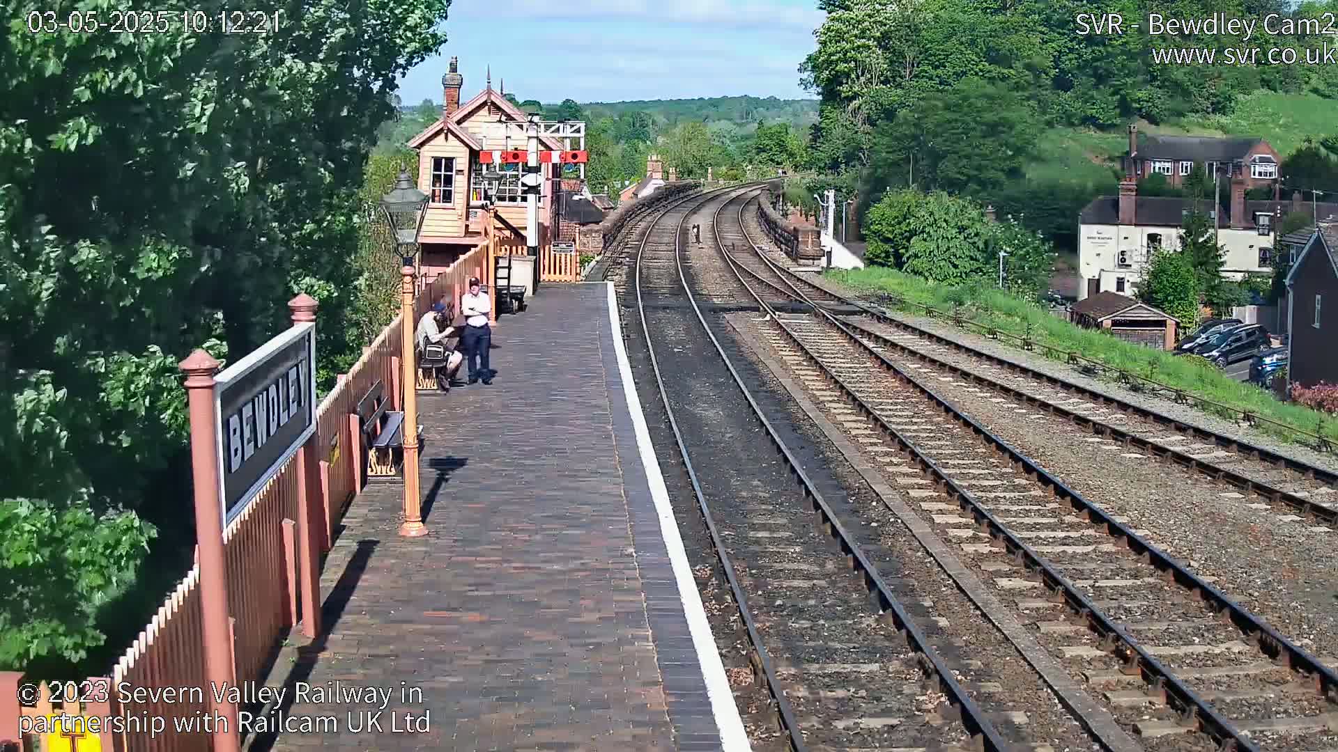 A sunny view of a train station platform with two people sitting, railway tracks extending into the distance, and a signal box.