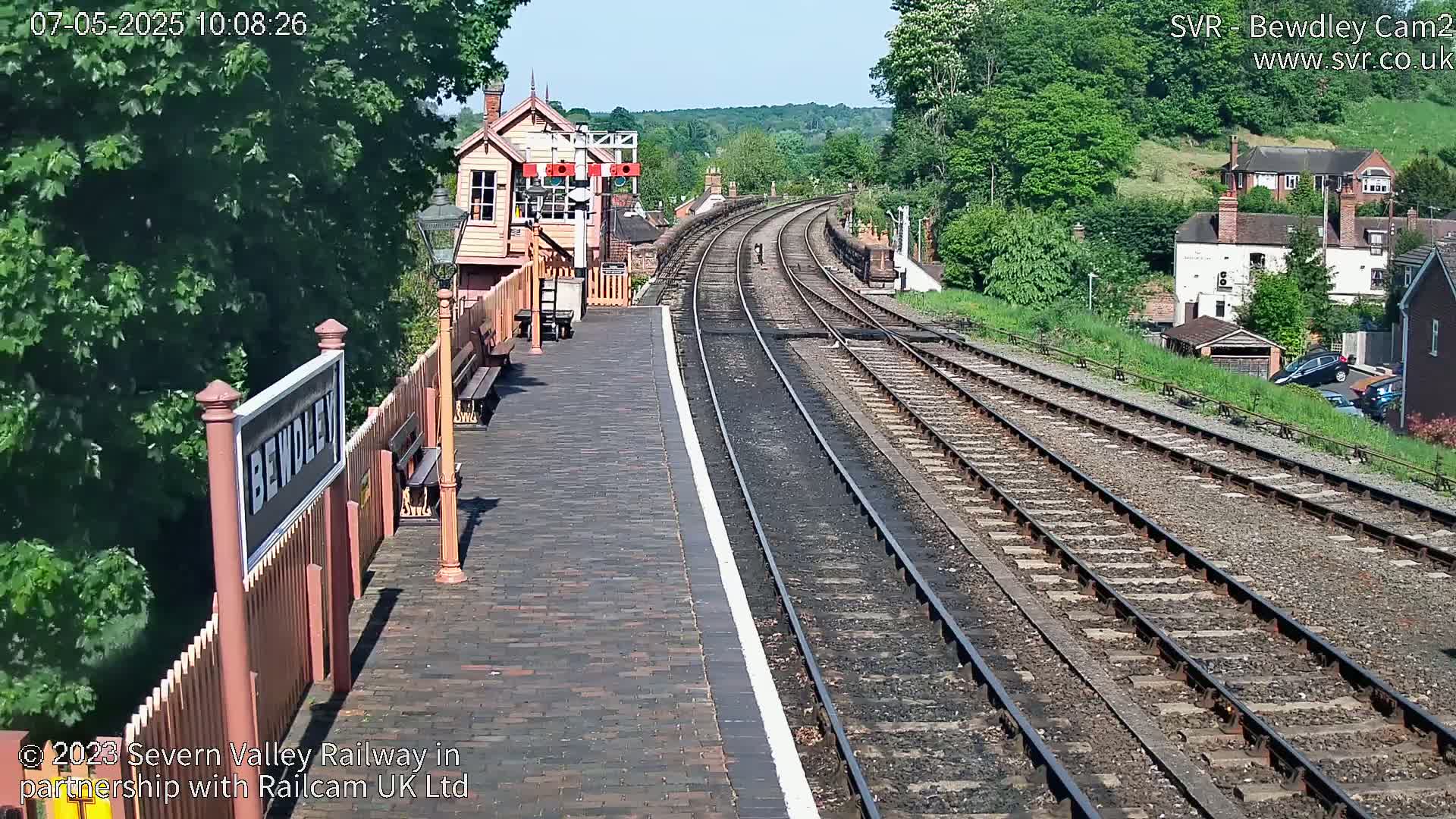 A brick train platform with a signal box and curving train tracks extends into a sunny, tree-lined distance.