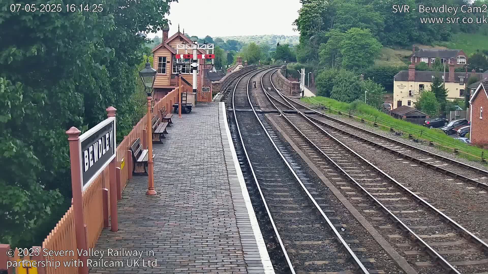 A train station platform with multiple train tracks stretches into the distance under an overcast sky, surrounded by lush green trees and buildings.